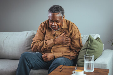 A senior black male appears distressed while clutching his chest, sitting on a grey sofa with a...