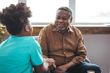 A mid-adult Black nurse attentively listens to her cheerful elderly patient in a well-lit home,...
