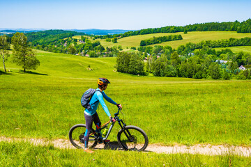 Woman with backpack riding mountain bike on rural road along green fields in Jizera Mountains, Singletrk pod Smrkem, Czech Republic