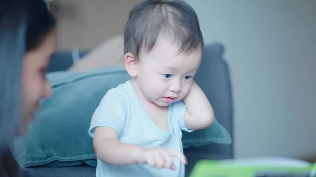Curious asian boy preschooler sitting next to the couch while listening to story from the book, young adult asian female reading book for her child, kids development skills, fast learner age