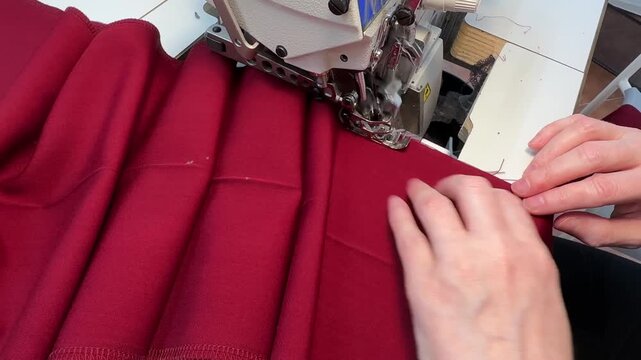 A close-up view of hands as they guide piece of red fabric through serger machine. machine as machine processes edge of the cut creating clean and professional finish fabric is folded over on itself.