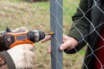 A man is building a fence at his dacha. Using an electric drill, he screws a metal mesh chain-link...