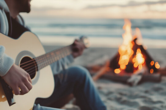 Portrait of happy smiling young man playing a guitar on a sea or ocean sand beach shore near the campfire bonfire. Evening sunset leisure, friends weekend enjoyment and relaxation,