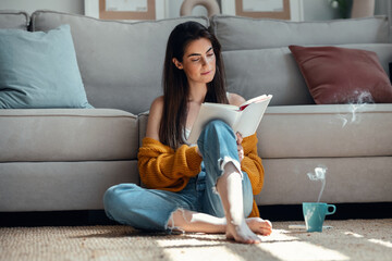 Pretty young woman reading a book and drinking a cup of coffee while sitting on the floor at home