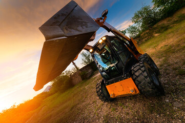 small bulldozer stands in green grass against the backdrop of sun rays in a forest belt at sunset © yanik88