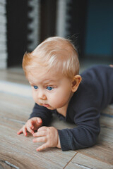 A blue-eyed baby lies on the floor and looks into the camera. A baby with white hair in a gray romper is playing at home. Clothes for babies.
