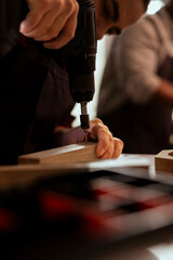 Carpenter drilling hole into timber block in studio helped by coworker, close up. Woodworking expert in joinery inserting nail into piece of wood using power drill with apprentice in background