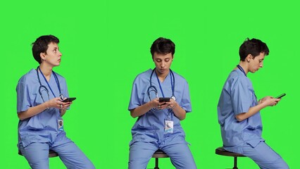 Obraz premium Nurse browsing online websites on smartphone app in studio, sitting on a chair against greenscreen backdrop. Medical specialist in scrubs using mobile phone and social media texting. Camera B.