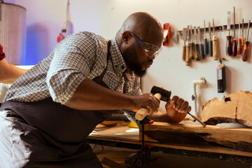 BIPOC man in carpentry shop wearing protection equipment while carving wood to prevent workplace accidents. Cabinetmaker equipped with safety goggles while using chisel and hammer to avoid injury