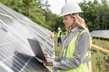 A woman wearing a yellow vest and a hard hat is looking at a laptop. She is working on a solar panel installation