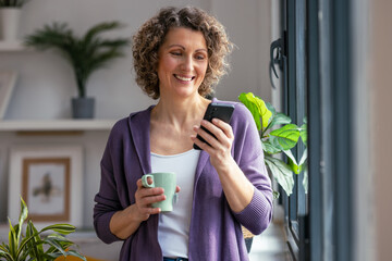 Pretty mature woman using her mobile phone while drinking coffee near to the window in the living...