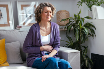 Unhealthy mature woman with stomachache leaning on the couch in the living room at home