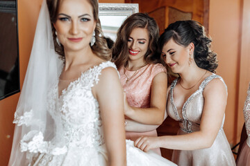 Three women are getting ready for a wedding. One of them is wearing a white dress and the other two are helping her with her dress