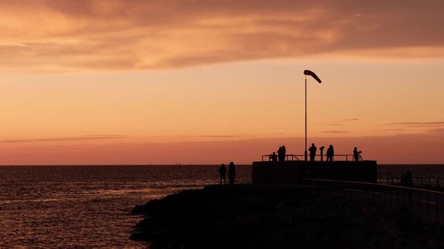 Unrecognisable people watching the sunset next to a windbag from a pier. Colourful lights above the reflecting sea surface, a seagull flying away. Summer evening at the Baltic Sea in Warnem&uuml;nde.