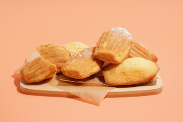 Wooden board with delicious madeleines and powdered sugar on orange background