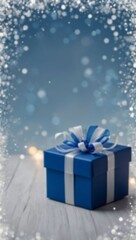 A blue gift box with a white bow sits on a white wooden surface with blurred lights and snowflakes in the background, out of focus, copy space