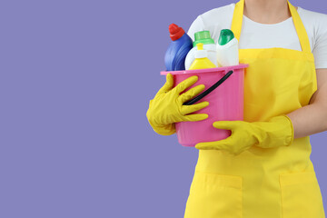 Female janitor with bucket of cleaning supplies on lilac background, closeup