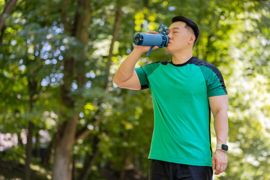 Sporty athletic thirsty guy Chinese man runner jogger drinking water after workout in park outdoors drink sport bottle, healthy habit active young Asian lifestyle refreshment hydration looking away.