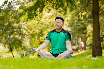 Young Asian man sitting on mat in lotus position, relaxing in city park on summer day. Sporty Japanese guy practicing yoga meditation, receives positive energy, healthy lifestyle. Active sportsman.