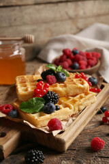 Board of sweet Belgian waffles with different fresh berries and jar of honey on wooden background