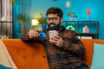Happy young Indian man doing payments online purchase shopping in internet store. Smiling Hispanic guy holding banking credit cards involved in verification process in mobile app on sofa at home room
