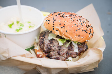 A take out hamburger with a side of potato salad.  Beef hamburger with lettuce and black sesame bun.