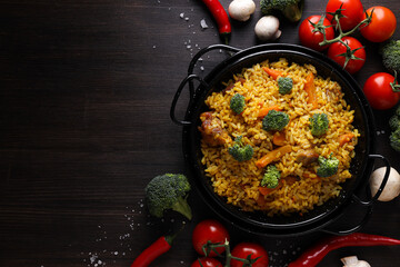 Pilaf in frying pan and vegetables on dark wooden background, space for text