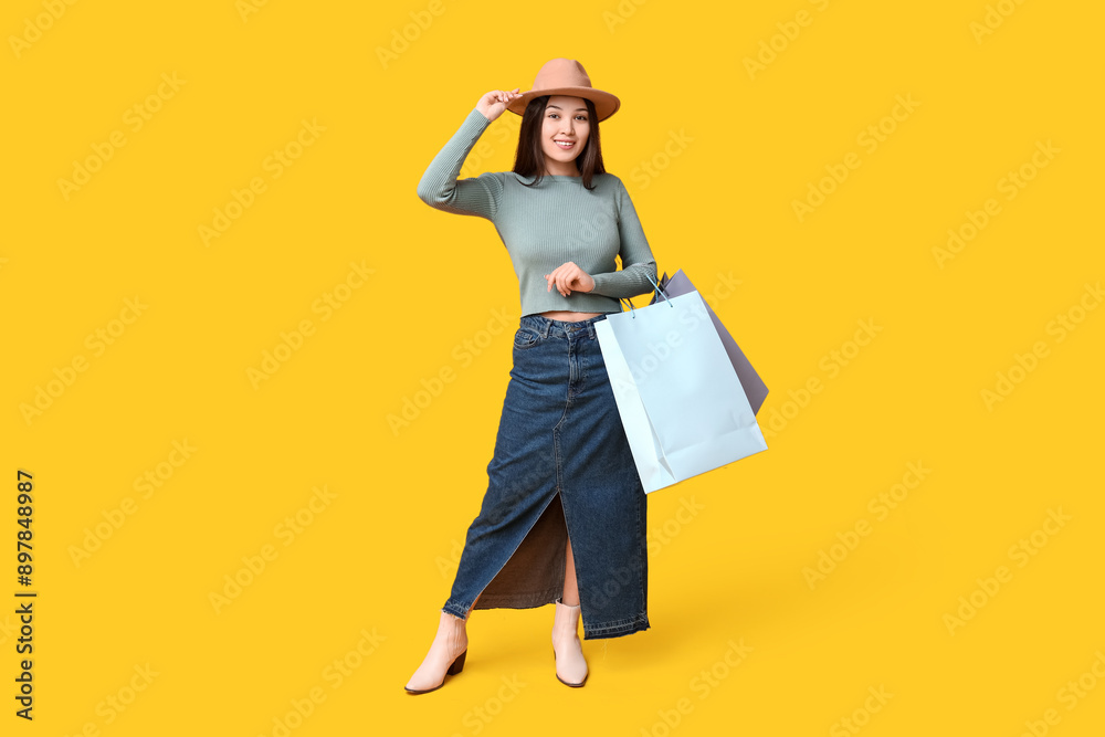 Sticker Fashionable young Asian woman in stylish denim skirt with shopping bags on yellow background