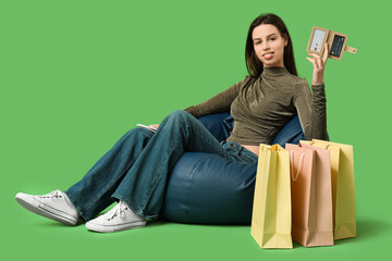 Young woman with credit card holder and shopping bags sitting on chair against green background