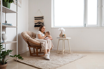 Beautiful young woman sitting in armchair with cup of tea in living room