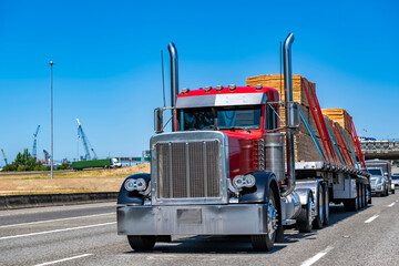Powerful classic red big rig semi truck tractor transporting fastened plywood on flat bed semi...