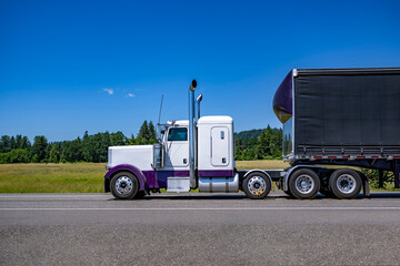 Profile of the classic white big rig semi truck with chrome exhaust pipes transporting cargo in black dry van semi trailer with front spoiler driving on the highway road