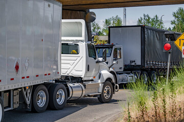 Two day cab white semi trucks with dry van and Conestoga  flat bed semi trailers meet on a narrow road under a railway bridge