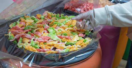 A gloved hand reaches into a clear plastic bag containing a large amount of colorful gummies. The gummies are piled high in a black bowl and are visible through the plastic bag.