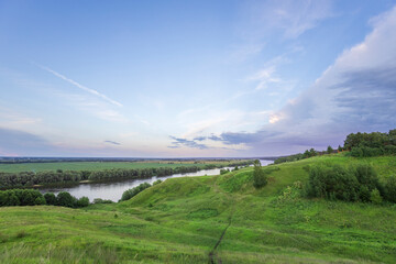 A winding river flows through a verdant valley, surrounded by lush green hills and trees. The sky...