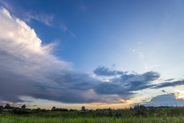 The sky above a lush field is filled with billowing clouds as the setting sun's warm glow hints at a tranquil evening in the distant village