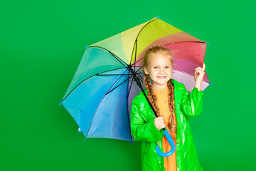 a child girl on an isolated green background with an umbrella and a raincoat smiles, space for text