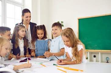 Friendly smiling female teacher is happy to teach fun lesson to children in elementary school. Cheerful schoolchildren with pencils and books are standing at one table and talking with teacher.