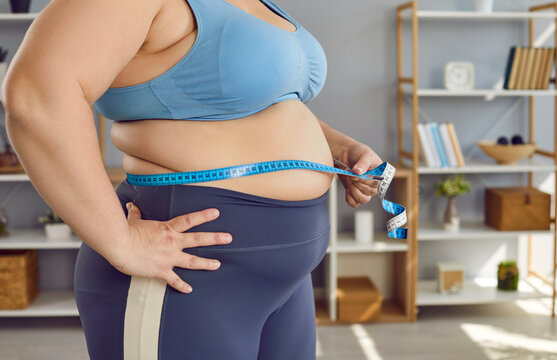 Crop shot overweight, obese woman in sports bra holding measuring tape around fat belly to check how much weight she has gained and how many centimeters she has added in her waist. Fat body concept