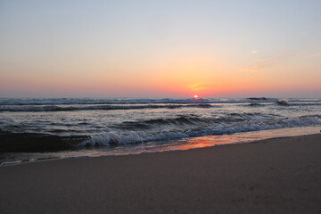 The sun sets behind the sea waves, its bright pink glow reflected on the surface of the water covering the dark sandy shore