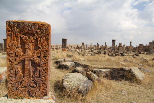 The image features a large number of khachkars, which are intricately carved stone crosses, standing in a field with a partially visible ancient church in the background under a cloudy sky.