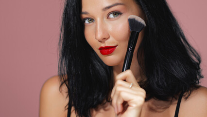 Glamorous Woman with Red Lips Applying Makeup Using Brush on Pink Background