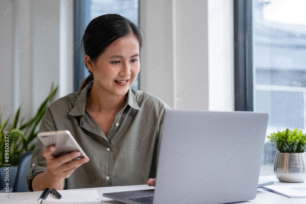 Wall mural Businesswoman sitting at desk on couch in workplace or at home working on laptop and analyzing data on charts and graphs and writing on papers to make business plan and strategies for company, 