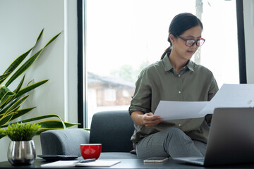 Businesswoman sitting at desk on couch in workplace or at home working on laptop and analyzing data on charts and graphs and writing on papers to make business plan and strategies for company, 