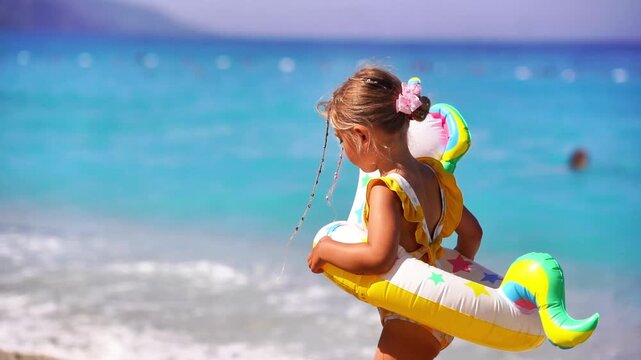 Summer vacation scene of a 5-year-old blonde girl walking on the beach with a swim ring around her waist, turquoise sea in the background. Capturing the joy and freedom of childhood during a sunny bea