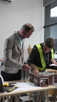 A Caucasian male engineer and an Italian male building contractor are planning and talking at a desk, reviewing house plans and a model of a house with wooden textures for renovation.