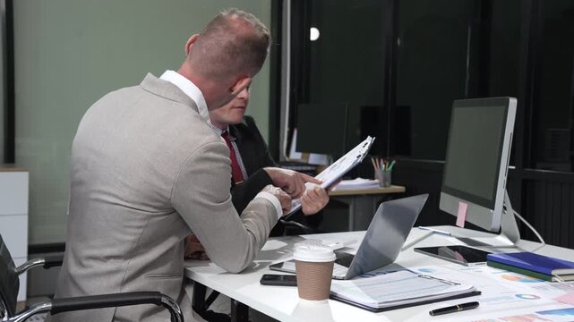 An Italian male and a Caucasian male, both in formal suits, sit at a business desk in an office meeting room with a desktop monitor and laptop, discussing business finance and investment strategies.