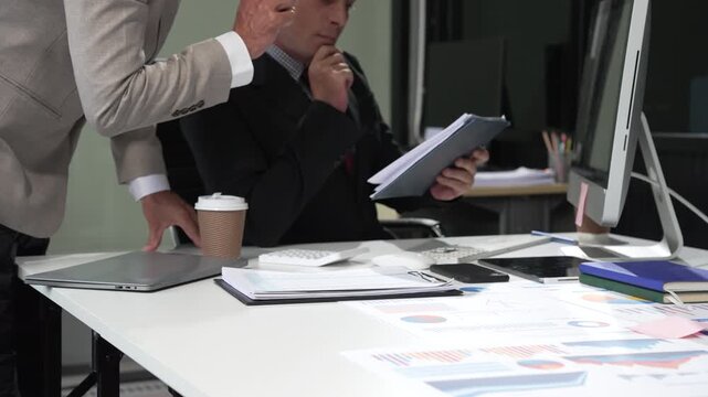 An Italian male and a Caucasian male, both in formal suits, sit at a business desk in an office meeting room with a desktop monitor and laptop, discussing business finance and investment strategies.