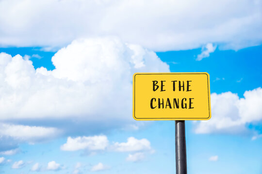 Yellow Sign with Inspirational Message "Be the Change" Against a Blue Sky with White Clouds
