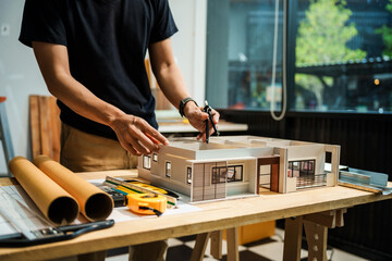 A man in a black t-shirt works at a desk, reviewing an architect's model of a house with various rooms, house plans, and wooden textures for a renovation project.